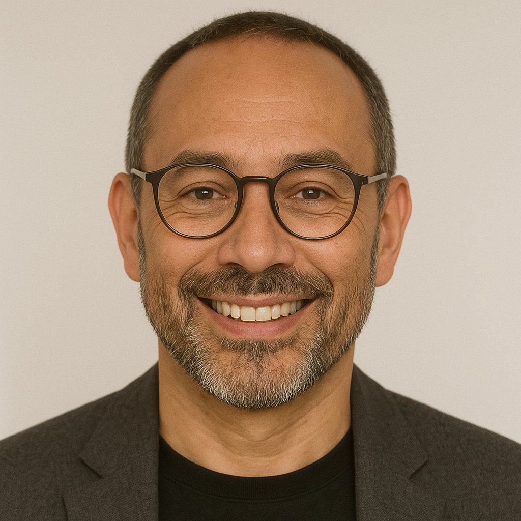 Homem sorridente de óculos e barba por fazer, em retrato profissional com blazer escuro e fundo neutro.